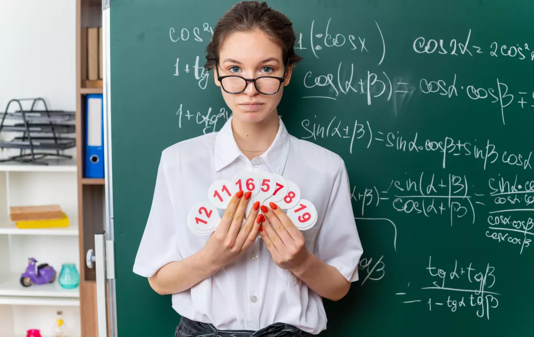 young-female-math-teacher-wearing-glasses-standing-front-chalkboard-holding-number-fans-looking-front-classroom