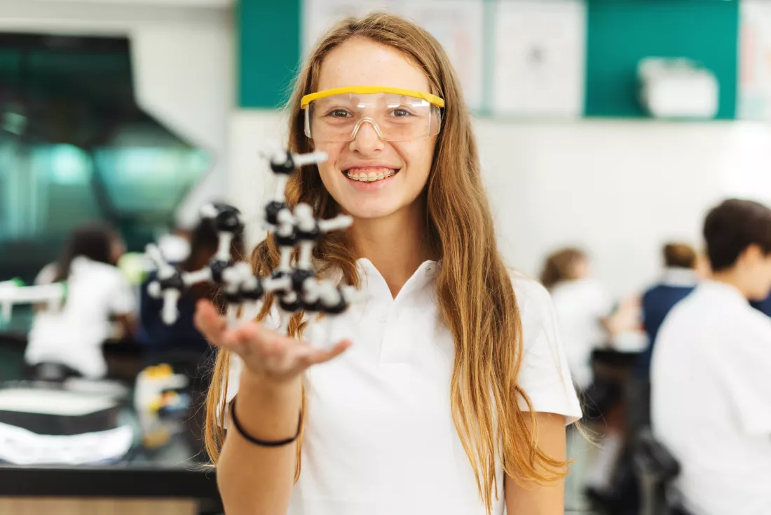 young-woman-science-lab-holding-molecular-model-wearing-safety-goggles-classroom-setting-with-students-background-focused-science-education-education-knowledge-science
